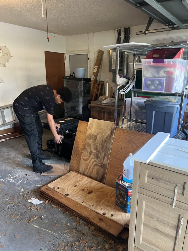Crew member bending over a black case beside plywood and garage storage shelves.
